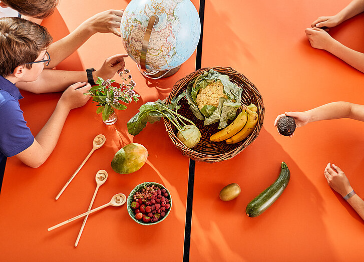 Auf einem orangenen Tisch steht ein Korb mit drei Bananen, einem Blumenkohl und einer Kohlrabi. Darum herum liegen außerdem eine Zucchini, eine Kiwi, eine Avocado, eine Mango und eine Schüssel mit Waldbeeren. Vier Jugendliche sitzen um den Tisch herum und betrachten die Lebensmittel sowie einen Globus, auf den zwei der Jugendlichen zeigen.